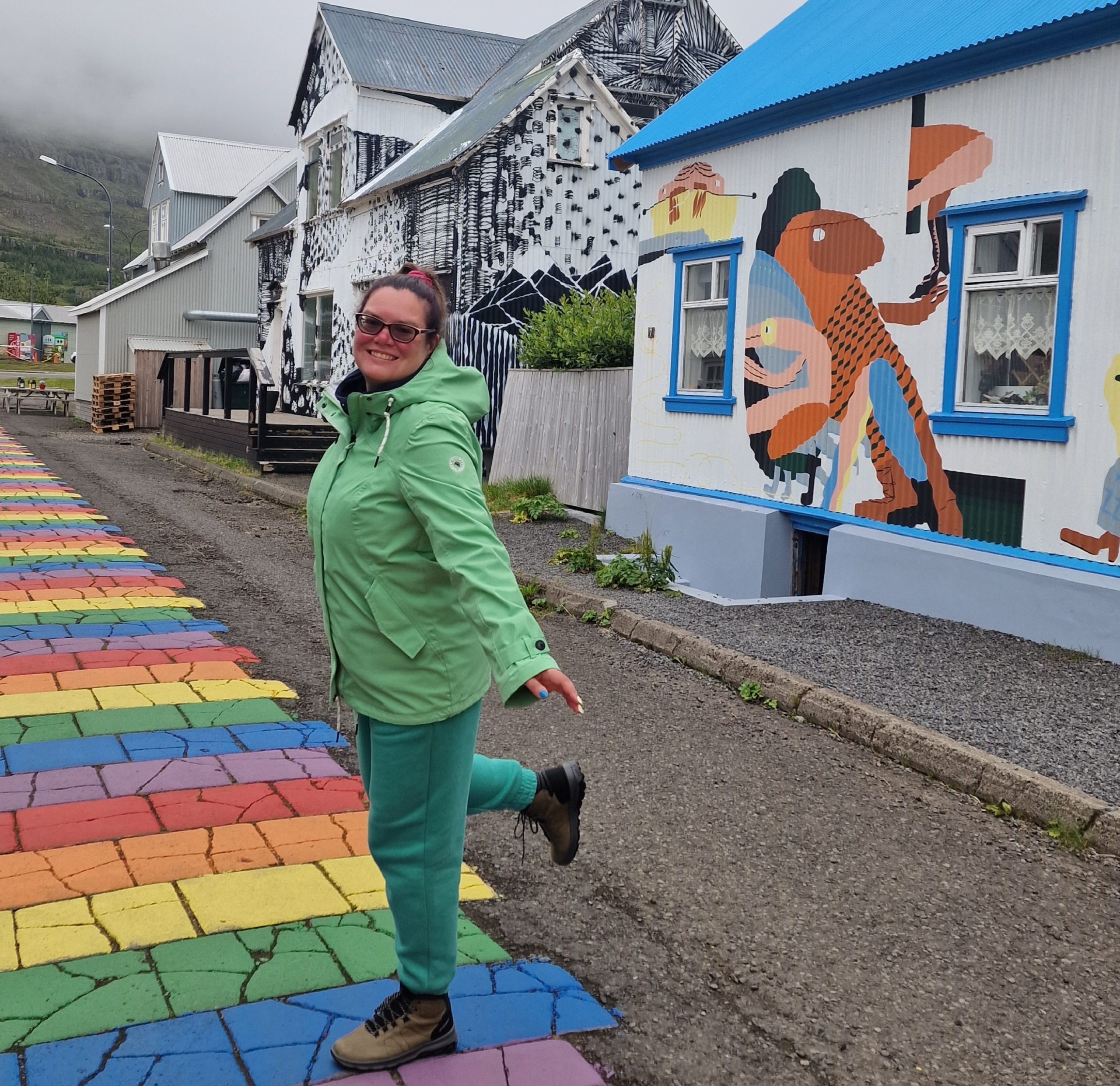 Ieva in Iceland - standing on a rainbow path with one leg raised up. Colorful buildings behind her.
