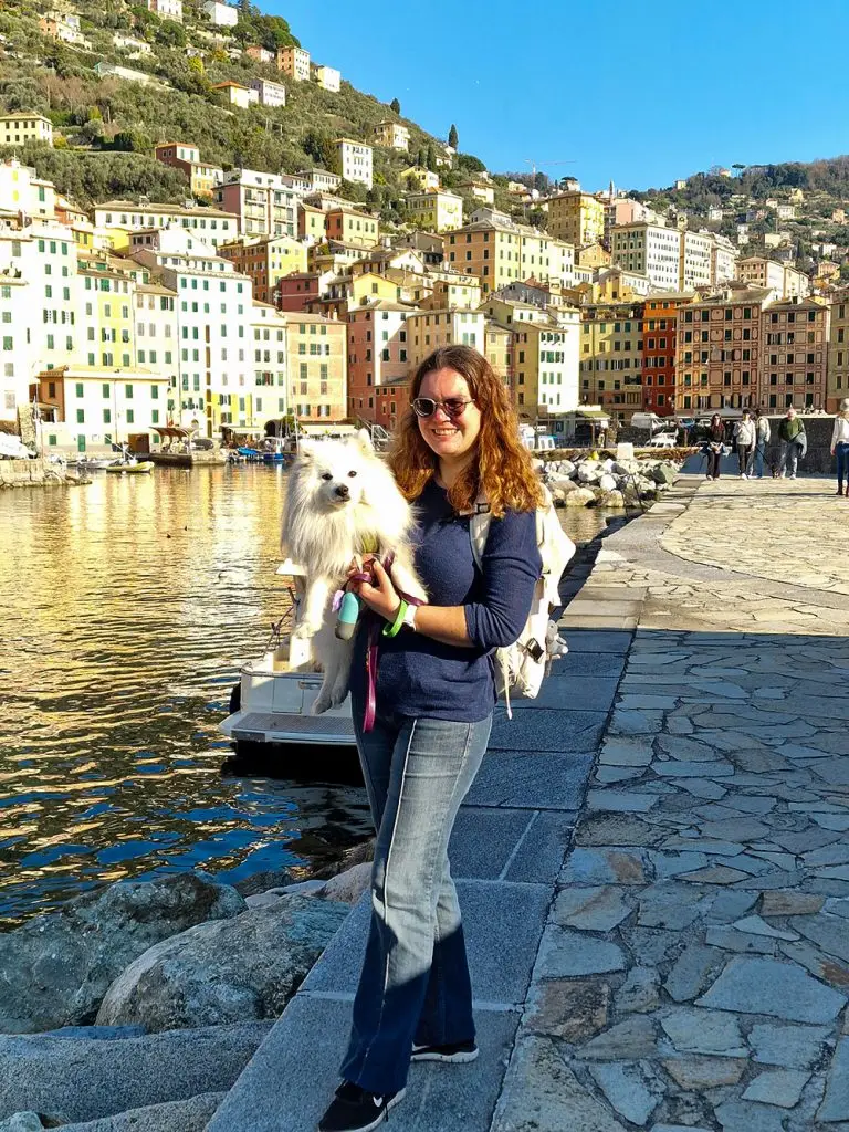A woman holding a white Japanese Spitz in her arms at Camogli harbor, with colorful waterfront houses, small boats, and people walking in the background.