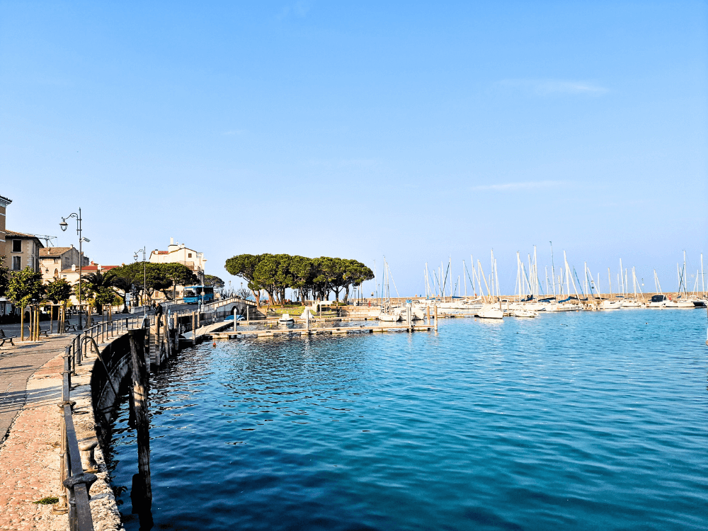 Where to stay on Lake Garda without a car: Peschiera or Desenzano? 7 Lakeside view in Desenzano del Garda with a quiet promenade, a few trees and sailboat masts visible in the distance.