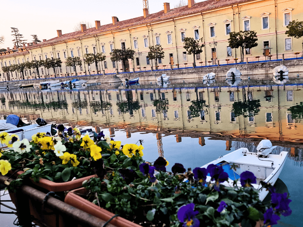 Where to stay on Lake Garda without a car: Peschiera or Desenzano? 5 Canal view in Peschiera del Garda with a historic military building, small trees along the wall, a moored boat and a large flower pot in the foreground.