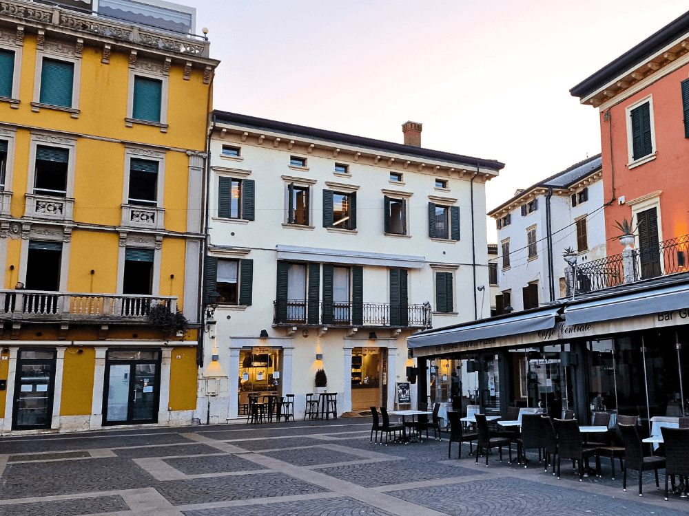 Where to stay on Lake Garda without a car: Peschiera or Desenzano? 2 A colourful square in Peschiera del Garda with pastel houses, a café terrace, tables and a sunshade.