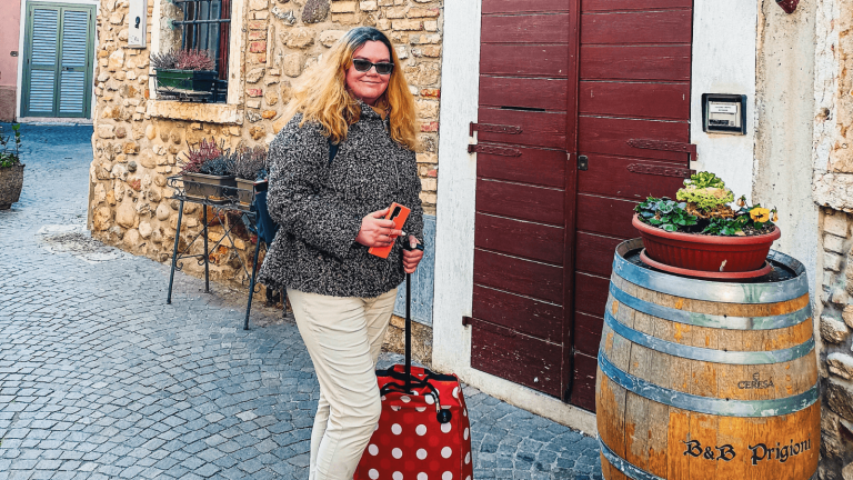 A woman standing with a suitcase in front of the B&B Prigioni entrance in Peschiera del Garda, holding her phone. A wooden barrel with the B&B name and a flower pot is placed by the door.