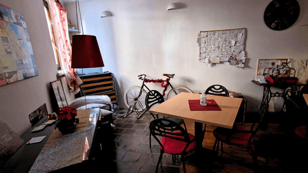 Where to stay on Lake Garda without a car: Peschiera or Desenzano? 3 A slightly messy, old-style Italian room at B&B Prigioni with a small table and chairs, a bicycle leaning against the wall, and three wall boards filled with guest notes and leaflets. Various items are scattered around the space.