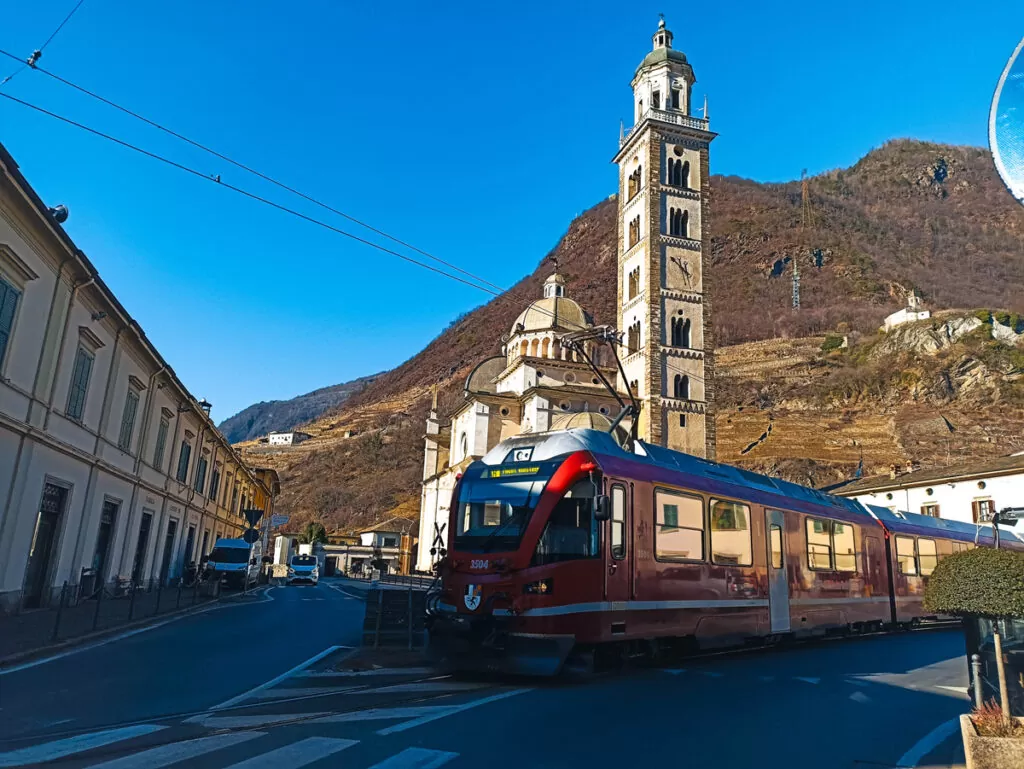 Bernina Express: a simple day trip into the Swiss Alps 1 The red Bernina Express travelling through a street in Tirano, with a church visible behind the train and tracks running along the road.