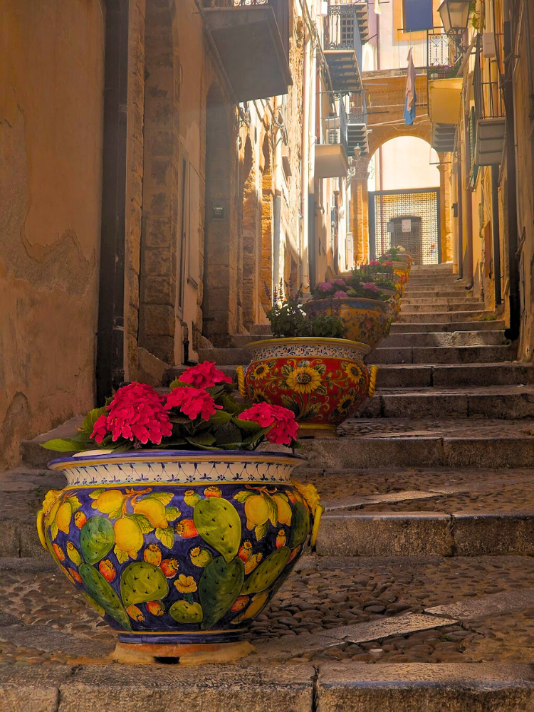 What to book first: flights or accommodation? 2 Narrow stone steps in Cefalù leading uphill between buildings, decorated with patterned plant pots filled with flowers.