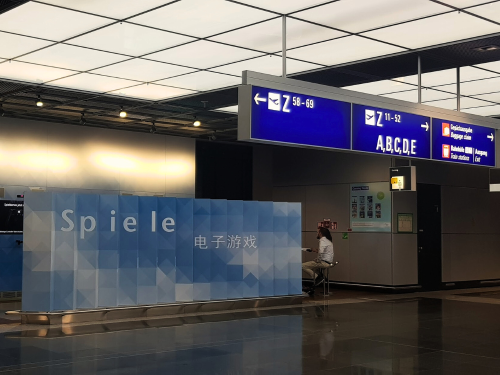 What to book first: flights or accommodation? 1 Interior of Frankfurt Airport with directional signs pointing to baggage claim and the train station, other signs showing gate directions, a person sitting on a chair, and the word “Spiele” written on a wall alongside Chinese characters.