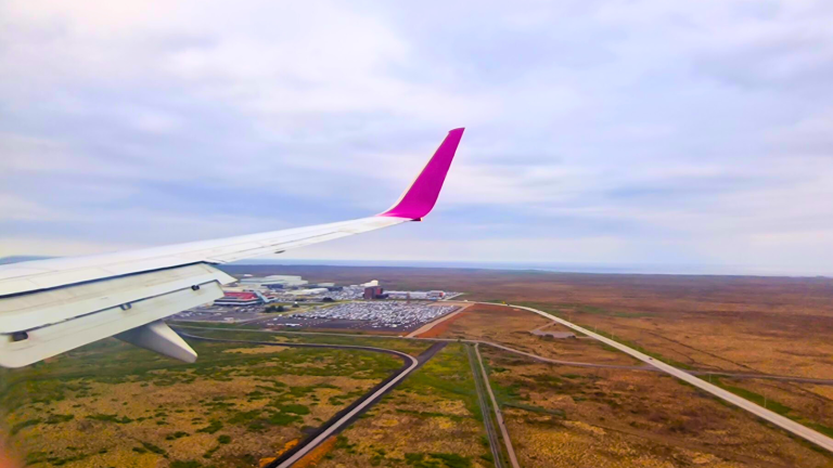White airplane wing with a pink tip flying low over fields, with a road and a few buildings visible below.