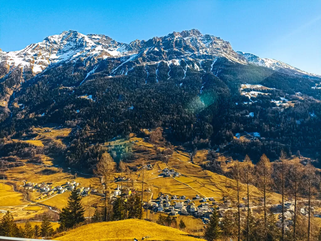 Bernina Express: a simple day trip into the Swiss Alps 6 View from the Bernina Express window showing snow-covered mountains, a yellowish meadow and a small village below.