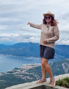 A woman wearing a hat stands on a cement wall, with one hand raised toward Kotor Bay, Montenegro.