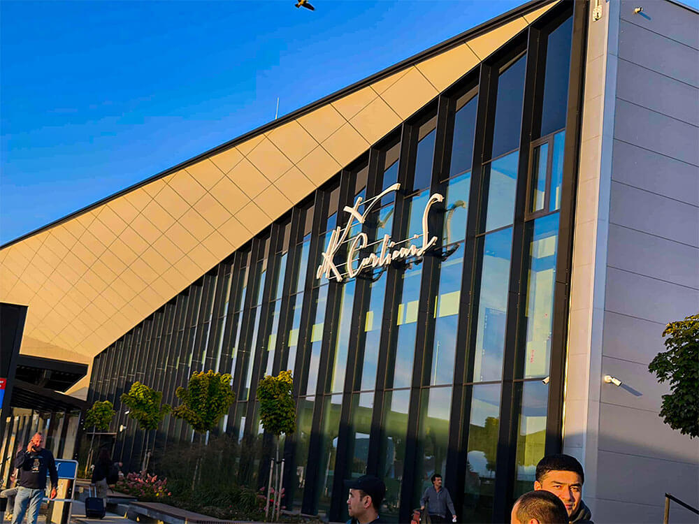 First time flying: what to do at the airport 2 The outside of a modern airport building is shown with a tall glass facade and an angled roofline. People stand near the entrance as warm sunlight hits the windows.