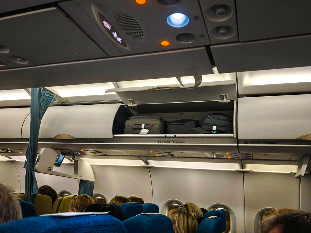 First time flying: what to do at the airport 8 Inside an aeroplane cabin the overhead lockers are open with bags stored inside. Passengers sit below while cabin lights glow along the ceiling.