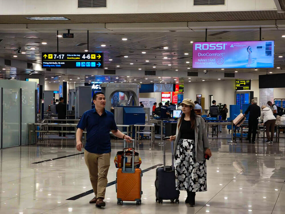 First time flying: what to do at the airport 12 Two travellers walk through a bright airport departures hall, each pulling a wheeled suitcase. Overhead signs point to boarding gates 7 to 17 and 4 to 6, with security screening equipment visible in the background.