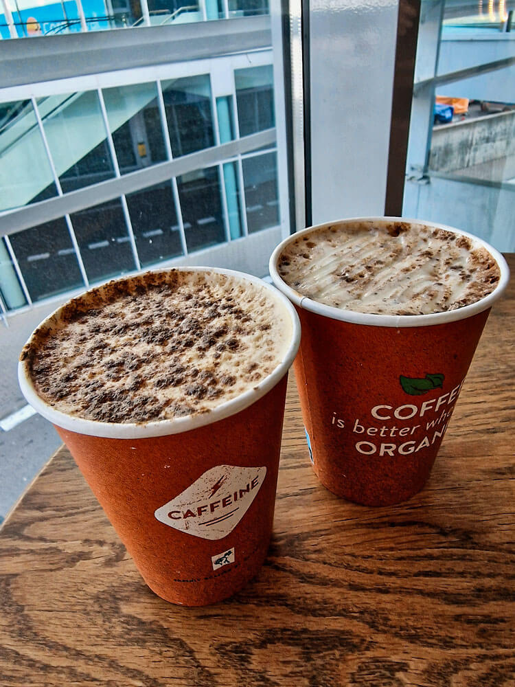 First time flying: what to do at the airport 6 Two takeaway cups of coffee sit on a wooden table by a window overlooking airport buildings. The foamy tops are dusted with cocoa or spice.