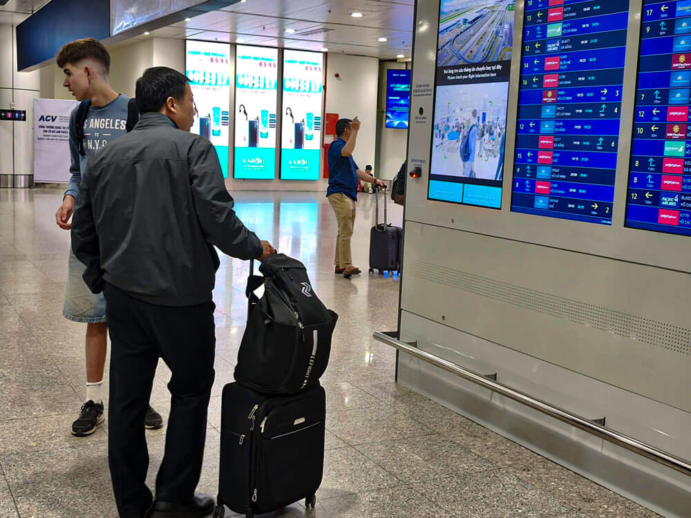 First time flying: what to do at the airport 3 Travellers stand in an airport terminal beside large flight information screens. A man pulls a wheeled suitcase while looking up at the departures board.
