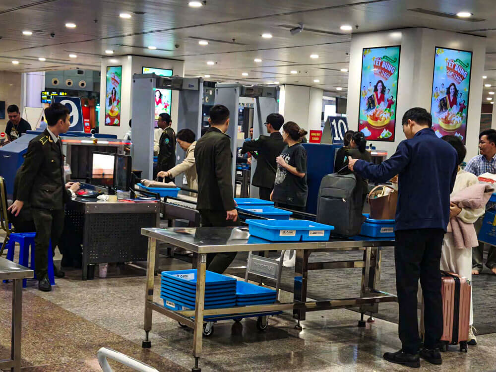 First time flying: what to do at the airport 4 Passengers queue at an airport security checkpoint, placing bags into blue trays on a metal table. Security staff stand beside the X ray machine and walk through metal detector frames while people prepare their belongings.