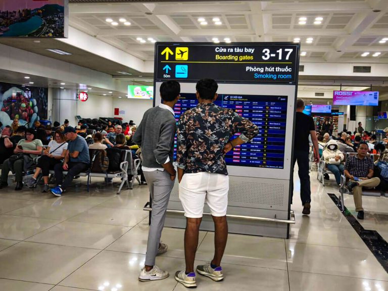 Two travelers stand in front of a flight information board in a busy airport terminal while other passengers sit and wait nearby. Overhead signs point to boarding gates 3 to 17 and a smoking room, helping orient anyone who is first time flying and figuring out where to go at the airport.