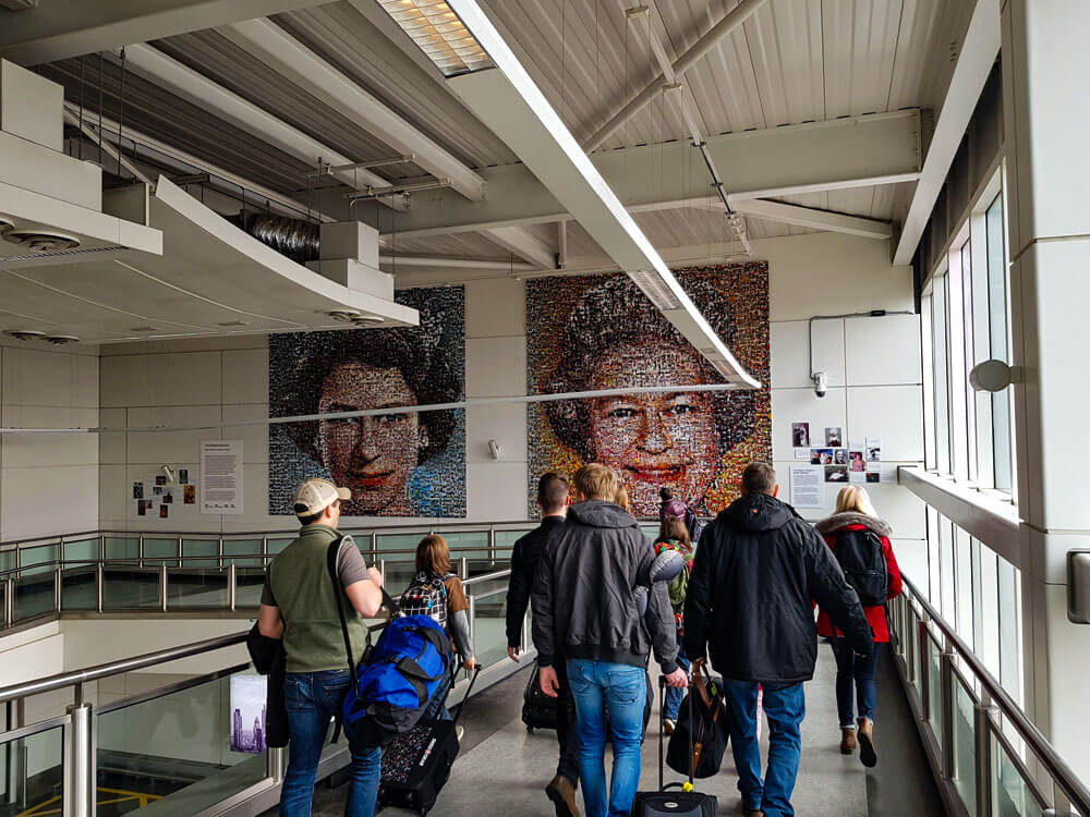 First time flying: what to do at the airport 10 Travellers with backpacks and wheeled luggage walk along an enclosed airport walkway. Large mosaic style portraits are displayed on the wall ahead.
