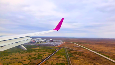 White airplane wing with a pink tip flying low over fields, with a road and a few buildings visible below.