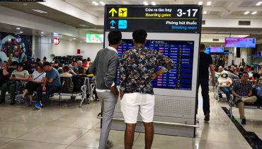 Two travelers stand in front of a flight information board in a busy airport terminal while other passengers sit and wait nearby. Overhead signs point to boarding gates 3 to 17 and a smoking room, helping orient anyone who is first time flying and figuring out where to go at the airport.