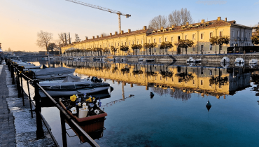A canal in Peschiera del Garda with a historic military building lined with small trees, several boats moored along the water and a construction crane visible behind the building, hinting at off-season renovation work.
