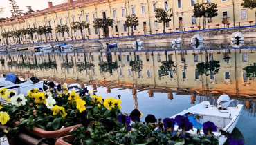Canal view in Peschiera del Garda with a historic military building, small trees along the wall, a moored boat and a large flower pot in the foreground.
