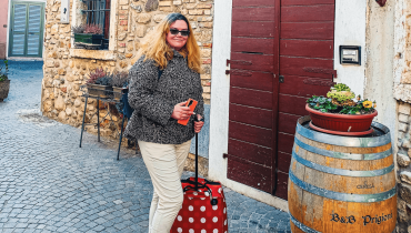 A woman standing with a suitcase in front of the B&B Prigioni entrance in Peschiera del Garda, holding her phone. A wooden barrel with the B&B name and a flower pot is placed by the door.